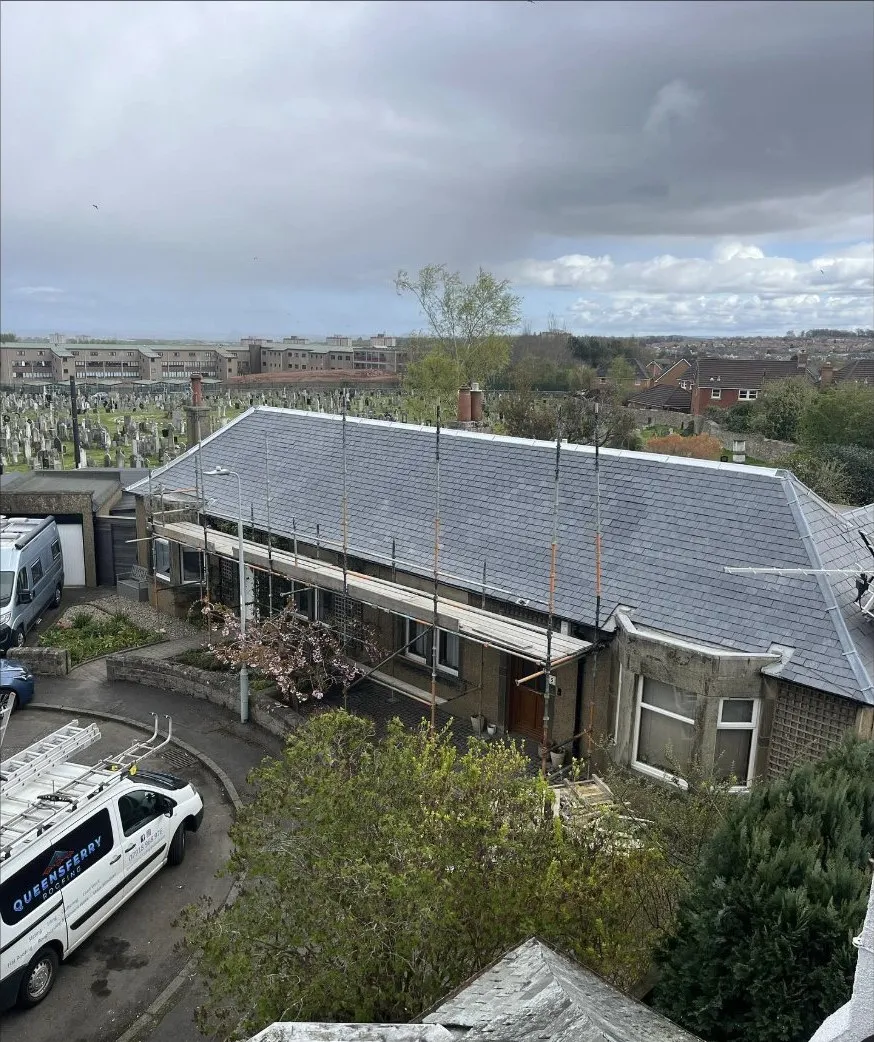 A completed slate re-roof shot from height, scaffolding still up, Edinburgh rooftops behind and the Queensferry Roofing van in the street below